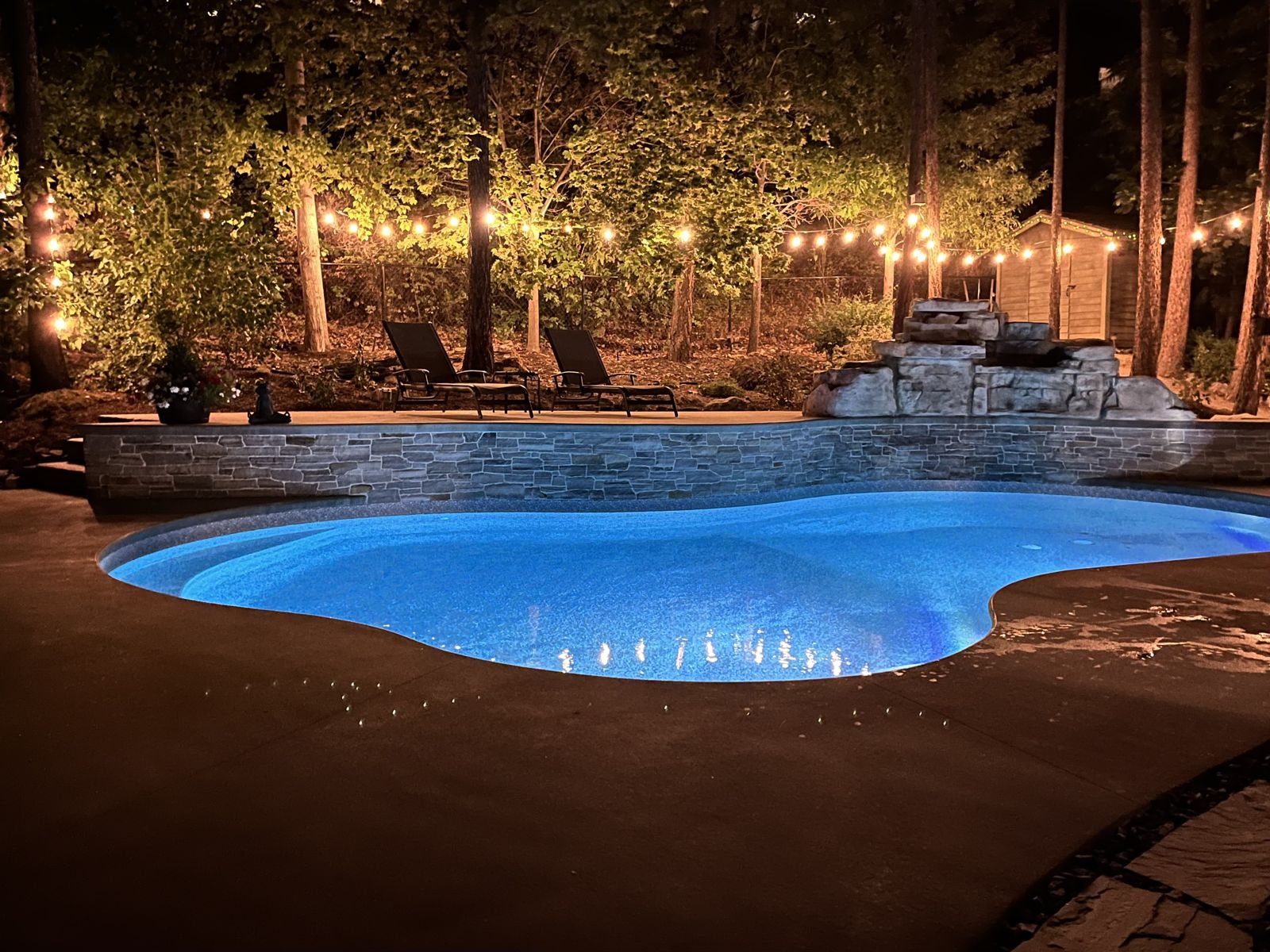 Freeform pool lit at night with a stone waterfall and string lights